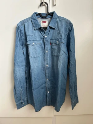 Blå klassisk jeansskjorta från Levi's - Säljer en klassisk blå jeansskjorta från Levi's med två bröstfickor och vita knappar framtill. Skjortan har lång ärm och en avslappnad passform. Perfekt till jeans eller kjol för en cool denimlook.