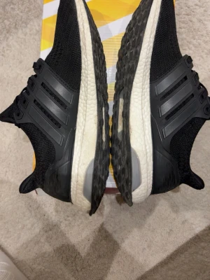 Svarta Adidas Ultraboost sneakers - Säljer ett par svarta Adidas Ultraboost sneakers med stickad ovandel och vita Boost-sulor. Skorna har klassiska tre ränder på sidan och en bekväm, responsiv sula som ger grym komfort. Perfekta för dig som vill ha både stil och funktion i vardagen.