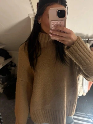 Beige oversized stickad tröja - Mysig beige stickad tröja med hög krage och breda ribbade muddar vid ärmslut och nederkant. Tröjan har en oversized passform och är perfekt för lager på lager. Materialet ser mjukt och fluffigt ut, vilket ger en avslappnad och trendig look.