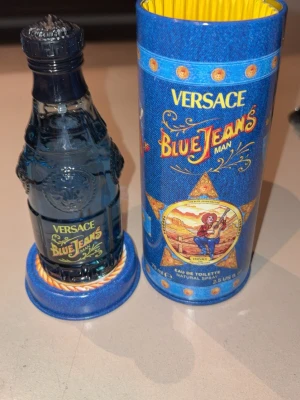 Versace Blue Jeans Man Eau de Toilette - Versace Blue Jeans Man Eau de Toilette i en blå glasflaska med detaljerad relief och gul text. Flaskan har en unik form och kommer med en färgstark, blå cylinderförpackning med denim-inspirerat tryck och illustration. Volymen är 75 ml.