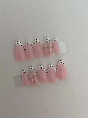Rosa press-on naglar med prickig spets - Snygga press-on naglar i ljusrosa med vit spets och svarta prickar. Några naglar har en 3D-blomma med gulddetalj för extra wow-känsla. Mandelformade och perfekta för dig som vill ha en trendig och söt look. Gjorda i plast.