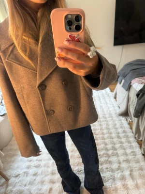 Kort brun dubbelknäppt rock - Snygg kort brun rock med dubbelknäppning och breda slag. Klassisk modell med rak passform och diskreta knappar framtill. Perfekt för dig som vill ha en stilren och trendig look. Materialet ser ut att vara ullblandning.