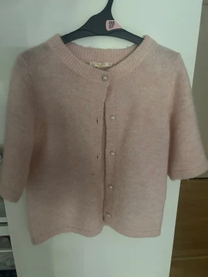 Ljusrosa kofta från Gina Tricot - Supersöt ljusrosa kofta från Gina Tricot med korta ärmar och pärlknappar framtill. Koftan är stickad i ett mjukt material och har en rund halsringning. Perfekt att slänga över en topp för en chill look.