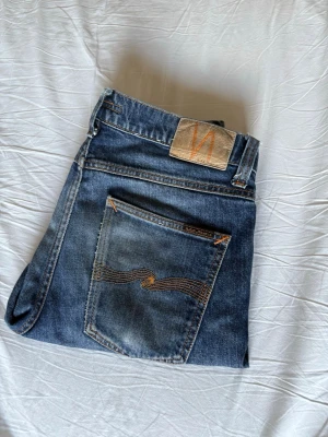 Blå jeans från Nudie Jeans - Säljer ett par klassiska blå jeans från Nudie Jeans med snygga slitningar och kontrastsömmar. Jeansen har en rak passform och bakficka med den ikoniska Nudie-bågen i orange. Perfekta för en avslappnad och trendig look.