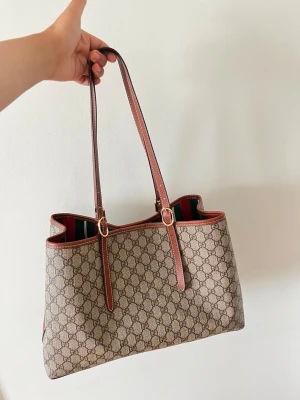 beige monogram handväska trendig - Snygg beige handväska från  med klassiskt monogrammönster och bruna läderdetaljer. Väskan har långa axelband och rödgrön randig insida. Perfekt för dig som vill ha en lyxig och rymlig väska i canvas och läder.