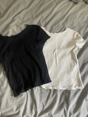 Ribbad svart och vit topp - Två ribbade toppar i svart och vitt med korta ärmar och lätt vågig kant vid hals och nederkant. Perfekta att matcha till jeans eller kjol för en enkel och clean look.