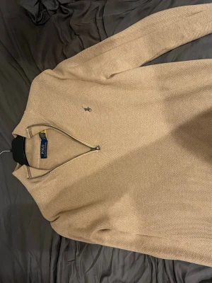 Beige halvzip från Polo Ralph Lauren - Snygg beige stickad tröja från Polo Ralph Lauren med halvzip och diskret logga på bröstet. Tröjan har lång ärm och ribbade muddar. Perfekt för dig som gillar klassisk och stilren design.
