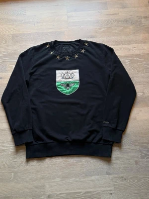 Svart Dolce&Gabbana tröja med broderad patch - Dolce & Gabbana svart tröja med broderad patch på bröstet som visar en krona och ett bi i vitt och grönt. Tröjan är XL men kan användas som (L o M) Runt halsen finns broderade guldstjärnor som ger en unik touch. Tröjan har rund hals, långa ärmar och ribbade muddar. 