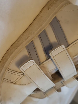 Vita Adidas sneakers med kardborre - Snygga vita sneakers från Adidas med klassiska tre ränder i ljusgrått på sidan och praktiska kardborreband. Skorna har en låg profil och detaljer i ljusgrått samt Adidas-logga bak på hälen. Perfekta för en clean och sportig look. obs! Storlek 26 (barn).