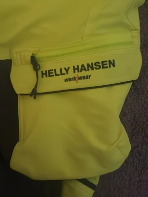 Gula arbetsbyxor Helly Hansen - Säljer ett par gula arbetsbyxor från Helly Hansen Workwear med flera praktiska fickor och svarta detaljer. Byxorna har dragkedja och knappar samt förstärkta partier på knäna. Perfekta för dig som vill ha funktionella och tåliga byxor.