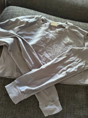 Ljusgrå cropped sweatshirt - Snygg ljusgrå cropped sweatshirt med långa ärmar och ribbade muddar. Tröjan har en enkel design med en liten vit logga på bröstet. Perfekt för träning eller chill dagar. Mjuk och bekväm bomullsblandning. Från gymshark
