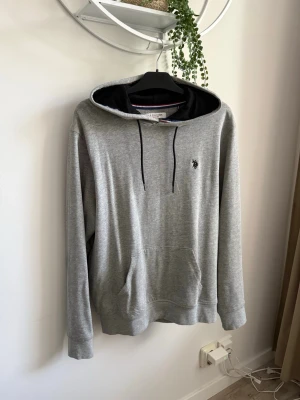 Grå hoodie med svart huva - Snygg grå hoodie med svart insida i huvan och svarta dragsnören. Klassisk känguruficka framtill och diskret broderad logga på bröstet. Perfekt för chill dagar eller när du vill ha en enkel och stilren look. ENDAST ANVÄND 1 GÅNG.