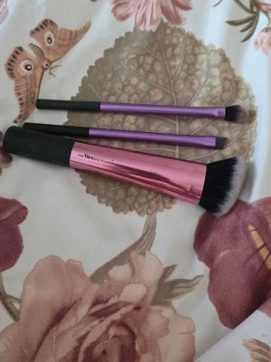 Sminkborstar från Real Techniques - Tre sminkborstar från Real Techniques. En större borste med rosa metallhandtag och två mindre borstar med lila handtag. Alla har svarta skaft och mjuka syntetiska borst. Perfekta för applicering av foundation, ögonskugga och detaljer.