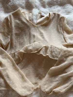Beige blus med volang och struktur - Supersöt beige blus med långärmad design och volangdetalj över bröstet. Tyget har en diskret prickig struktur som ger en trendig känsla. Perfekt för dig som gillar romantiska och stilrena plagg. Blusen har rund halsringning och är lätt att matcha.
