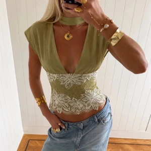 Grön croppad topp med spetsdetaljer - Trendig grön croppad topp med djup v-ringning och breda axlar. Toppen har vita spetsdetaljer med blommigt mönster i midjan och en matchande choker runt halsen. Perfekt för dig som vill sticka ut med en unik och snygg stil.