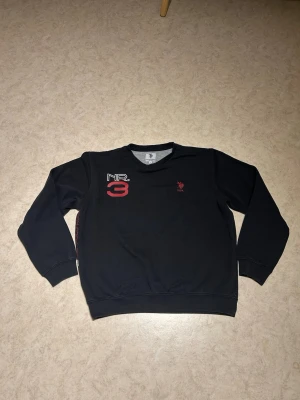 Svart stickad tröja från US Polo Assn - Svart stickad tröja från U.S. Polo Assn med rund halsringning. Tryck med 'NR. 3' i vitt och rött på bröstet samt röd USPA-logga. Långa ärmar och ribbade muddar vid ärmslut och nederkant. Perfekt för en sportig och avslappnad stil.