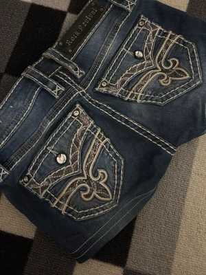 Blå jeansshorts med broderade fickor - Säljer ett par blå jeansshorts med snygga broderade detaljer och knappar på bakfickorna. Shortsen har kontrastsömmar och coola mönster på ficklocken. Perfekta för dig som vill sticka ut lite extra i sommar!