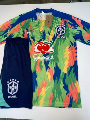 Färgglatt Brasilien fotbollsställ Nike - Säljer ett Brasilien fotbollsställ från Nike med färgstarkt mönster i grönt, blått, gult och orange. Tröjan har CBF-logga, sponsortryck och korta ärmar. Matchande blå shorts med CBF-logga ingår. Perfekt för dig som vill sticka ut på planen!