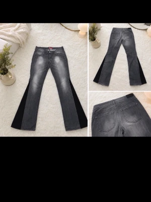 Gråa utsvängda jeans med svarta paneler - Säljer ett par gråa jeans med breda svarta paneler längs benen. Jeansen har utsvängda ben och klassisk femficksdesign. Perfekta för dig som vill sticka ut med en cool och unik look. STORLEK XL MEN PASSAR SOM storlek L. Har en liten fläck på änden av jeansen men går bort i tvätten