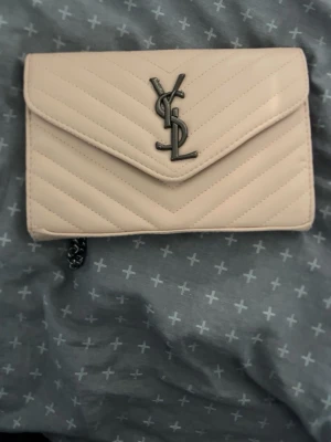 Beige kuvertväska med kedja YSL - Snygg beige kuvertväska från YSL med quiltat mönster och silverfärgad kedja. Väskan har ett stort YSL-emblem framtill och stängs med lock. Perfekt för dig som vill ha en stilren och trendig accessoar.