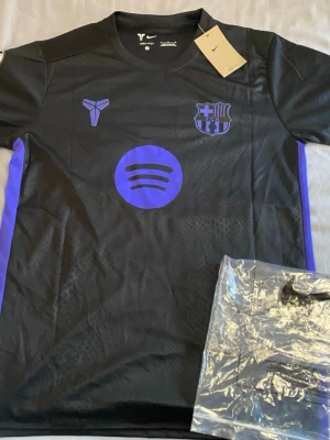 Svart FC Barcelona tröja Nike - Snygg svart FC Barcelona matchtröja från Nike med lila detaljer och tryck. Tröjan har klubbmärke, sponsorlogga och Kobe Bryant-symbol på bröstet samt Lamine Yamal och nummer 10 på ryggen. Tillverkad i lätt och ventilerande material, perfekt för fotboll eller träning.