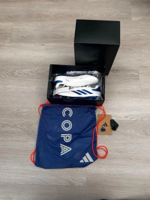 Adidas Copa fotbollsskor vita/blå - Säljer ett par Adidas Copa fotbollsskor i vitt med blå detaljer och röda logotyper. Skorna har klassisk snörning, tre blå ränder på sidan och är tillverkade i syntetmaterial. Levereras med blå förvaringspåse och extra tillbehör.