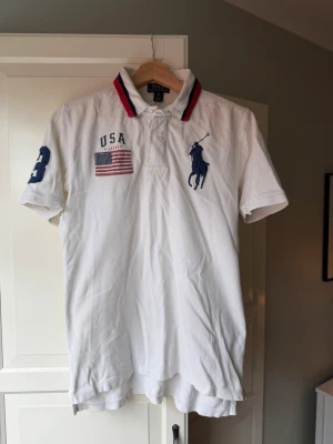Ralph Lauren piké  - Storlek jr:XL som motsvarar ca XS i herrstorlek. Vit pikétröja från Ralph Lauren med klassisk krage och knappar. Stora blå loggan på bröstet, USA-flagga, och siffran 3 på ärmen. Kragen har röda och marinblå detaljer. Perfekt för en sportig och stilren look.