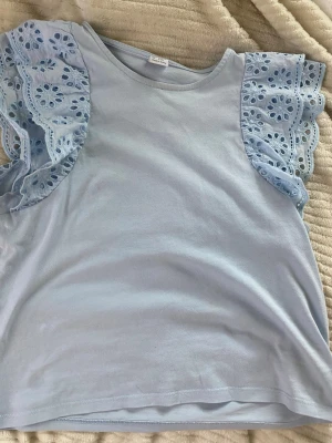 Ljusblå topp med spetsvolang - Supersöt ljusblå topp med broderade spetsvolanger på ärmarna. Toppen har rund halsringning och är tillverkad i mjuk bomull. Perfekt för dig som vill ha en feminin och fräsch look med extra detaljer på ärmarna.
