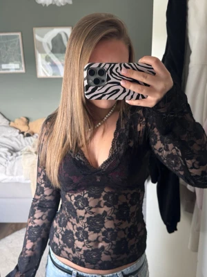 Svart spetsblus med v-ringning - Säljer en svart, transparent spetsblus med v-ringning och lång ärm. Blusen har ett blommigt spetsmönster och sitter tight mot kroppen. Perfekt att styla med ett linne eller bralette under för en trendig look.