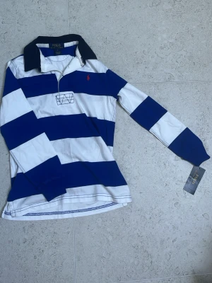 Randig rugbytröja från Polo Ralph Lauren barn - Säljer en klassisk långärmad rugbytröja från Polo Ralph Lauren med breda blå och vita ränder, mörkblå krage och röd logga på bröstet. Tröjan har knappar i halsen och är gjord i mjuk bomull. Perfekt för en sportig och avslappnad stil. Passar perfekt för sjuåringar.