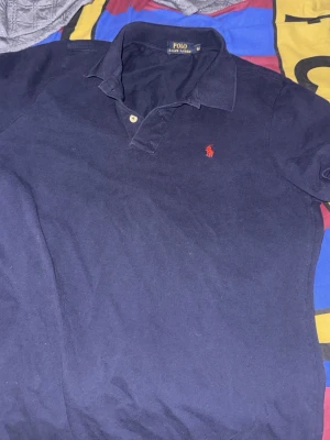 Marinblå pikétröja från Polo Ralph Lauren - Klassisk marinblå pikétröja från Polo Ralph Lauren med krage, två knappar och den ikoniska röda logotypen broderad på bröstet. Tillverkad i mjuk bomull som känns skön mot huden. Perfekt för en stilren och avslappnad look.