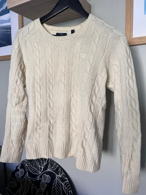 Kabelstickad offwhite tröja från gant  - Kabelstickad tröja i offwhite med rund halsringning och långa ärmar. Tröjan har ett klassiskt flätmönster och ribbade muddar vid ärmslut och nederkant. Perfekt för lager på lager och riktigt mysig till höst och vinter.