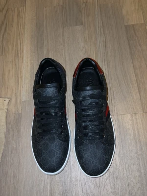 Svarta Gucci sneakers med röd/blå detalj - Snygga svarta sneakers från Gucci med monogrammönster, vita sulor och klassiska röd-blå ränder på sidan. Baksidan har metallicdetaljer i rött och blått. Skorna är låga och har svarta skosnören. Perfekta för dig som vill ha en exklusiv touch på din outfit.