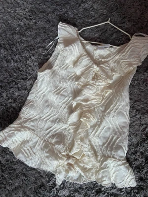 Vit volangblus med struktur - Supersöt vit blus med volangdetaljer framtill och ett strukturerat, lite skrynkligt tyg. Ärmlös modell med v-ringning. Från Gina tricot i storlek 164