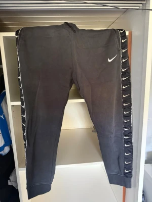 Svarta Nike mjukisbyxor med logga - Säljer ett par svarta mjukisbyxor från Nike med vita loggor längs sidorna och en större logga på låret. Byxorna har muddar vid bensluten och är gjorda i mjukt bomullsmaterial. Perfekta för chill dagar eller träning.