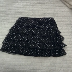 Svart volangkjol med vita prickar  - Säljer en svart kortkjol med vita prickar och flera lager volanger. Kjolen har en söt och lekfull look med sitt prickiga mönster och volangdetaljer. Perfekt för dig som vill sticka ut lite extra! Aldrig använd💕