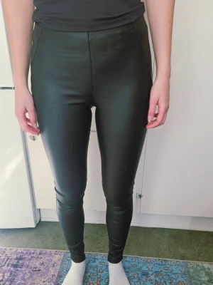 Svarta leggings i fuskskinn - Säljer ett par svarta leggings i fuskskinn med hög midja och tight passform. De har en slät framsida utan synliga fickor eller dragkedjor och ger en cool, stilren look. Perfekta att matcha med oversized tröjor eller crop tops. Ej retur.