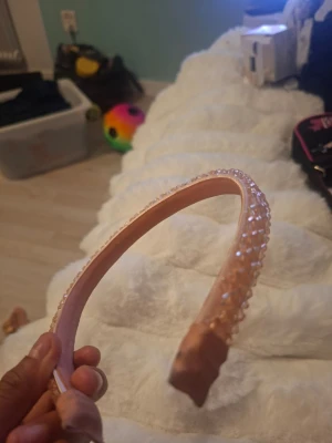 Rosa diadem med pärlband - Snyggt rosa diadem med ett band av små glittrande pärlor längs ovansidan. Diademet har en mjuk, vadderad insida och är täckt av ett satinliknande tyg. Perfekt accessoar för att lyfta din look med lite extra bling.