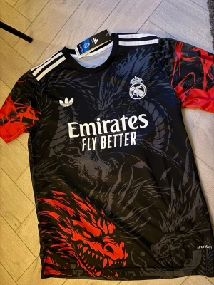 Real Madrid specialtröja – Ny med tags -   Säljer en snygg Real Madrid-tröja med unikt drakmotiv i svart och rött. Helt ny med tags kvar. Bekväm passform och perfekt för både fotboll och vardagsbruk.  