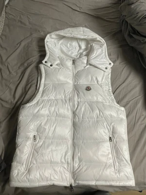 Vit dunväst med huva från Moncler - Snygg vit dunväst från Moncler med huva och två dragkedjefickor framtill. Västen har en glansig finish, Moncler-logga på bröstet och stängs med dragkedja. Perfekt för lager-på-lager och streetstyle.
