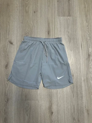 Grå träningsshorts från Nike - Snygga grå träningsshorts från Nike med elastisk midja och snörning. Shortsen har en loose fit och är tillverkade i ett lätt och snabbtorkande material, perfekta för träning eller chill. Klassisk Nike-logga på benet.