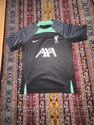Liverpool FC svart tränings t-shirt - Snygg svart tränings t-shirt från Nike med Liverpool FC-logga och AXA sponsortryck på bröstet. Tröjan har mörkgrå ärmar och gröna detaljer vid hals och sidor. Perfekt för fotbollsträning eller gymmet. Tillverkad i lätt och funktionellt material.
