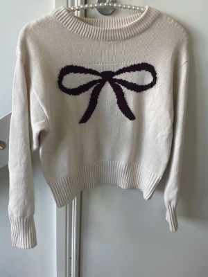 Vit stickad tröja med rosett - Söt vit stickad tröja med en stor svart rosett på bröstet. Tröjan har rund halsringning, långa ärmar och ribbade muddar vid ärmslut och nederkant. Perfekt för dig som gillar gulliga detaljer och enkel stil.