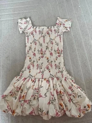 Blommig kortklänning med volang - Supersöt kortklänning i ljus färg med blommigt mönster. Klänningen har smockad överdel, volangig kjol och korta ärmar. Perfekt för sommarens alla tillfällen och ger en romantisk vibe.