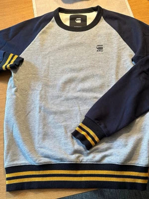 G-Star RAW sweatshirt blå/grå storlek S - Snygg sweatshirt från G-Star RAW med grå framsida och mörkblå ärmar. Tröjan har ribbade muddar och nederkant med gula och svarta ränder. Liten RAW-logga på bröstet. Perfekt för en avslappnad streetwear-look.