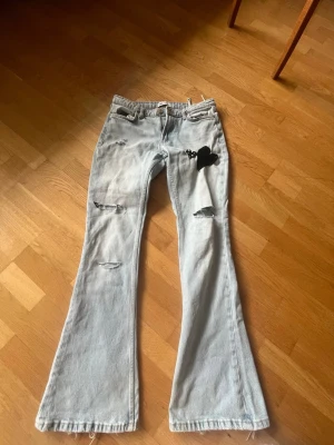 Ljusblå utsvängda jeans - Säljer ett par ljusblå jeans med utsvängda ben och slitna detaljer. Jeansen har en svart hjärtformad lapp på ena låret 