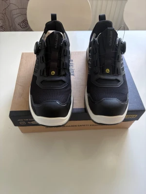 Blåkläder skor  - Snygga svarta sneakers med vit sula och smart snörningssystem med ratt. Ovandelen är i mesh och syntetmaterial för bra andningsförmåga och komfort. Tjock yttersula med bra grepp och detaljer i svart och grått. Perfekta för en sportig och modern look.