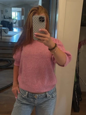 Rosa stickad tröja med puffärm - Söt rosa stickad tröja med korta puffärmar och ribbade muddar. Tröjan har en avslappnad passform och är gjord i ett mjukt, fluffigt material som känns skönt mot huden. Perfekt att styla med jeans för en trendig look. OBS: finns ett märke som inte går bort på tröjan! 
