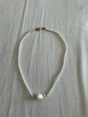 Vitt pärlhalsband med stor barockpärla - Säljer ett stilrent halsband med en stor barockpärla i mitten. Halsbandet har ett guldfärgat lås och är perfekt till sommaren.