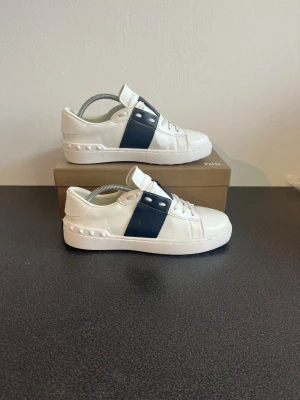 Vita sneakers Valentino Garavani - Snygga vita sneakers med den ikoniska mörkblåa breda randen.  Skorna har vita snören och en chunky sula med rutmönster undertill. 